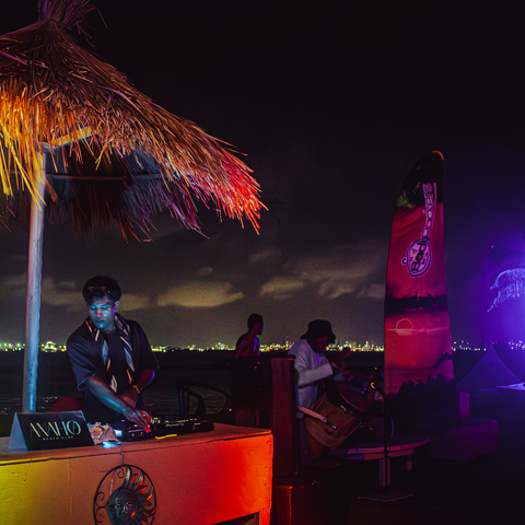 DJ at a tropical outdoor event with colorful lighting and a thatched roof.