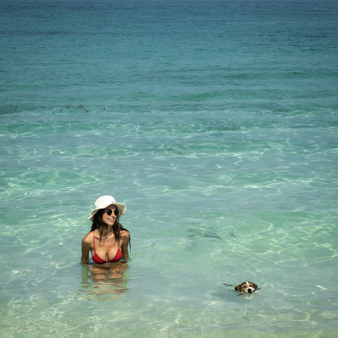 Woman in a bikini and hat standing in clear ocean water with a dog nearby.